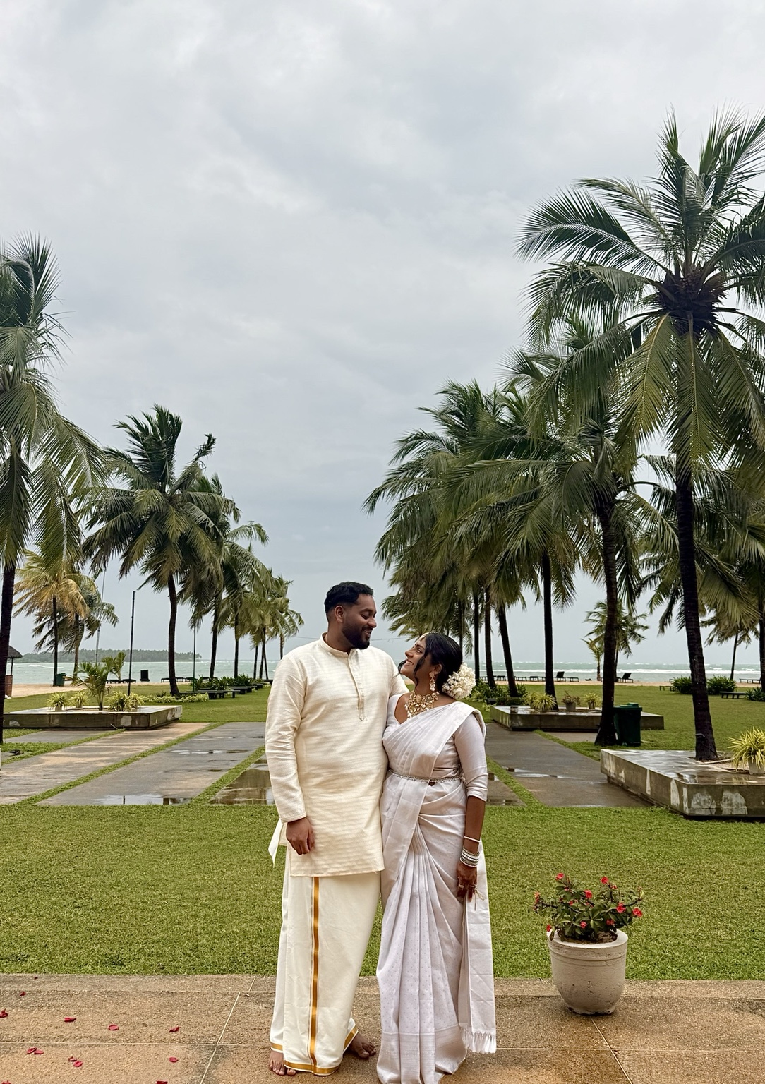 Aaranan and Aniththa in ivory traditional outfits, smiling together beneath coastal palms